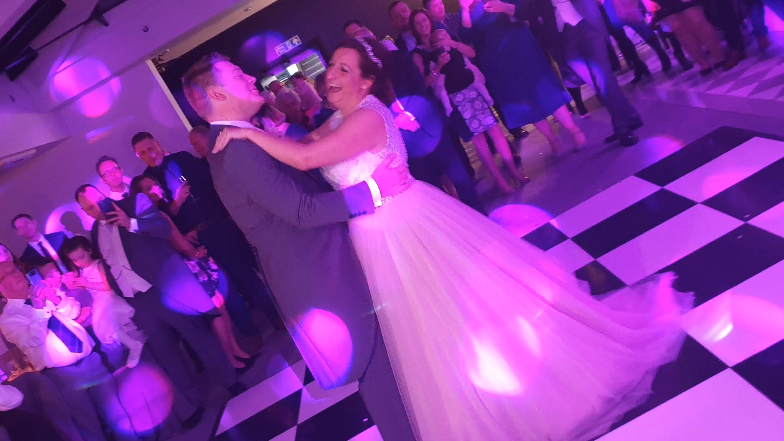 Bride and groom laughing during their first dance on a black and white checkered dance floor at Swynford Manor in Cambridgeshire, with pink DJ uplighting