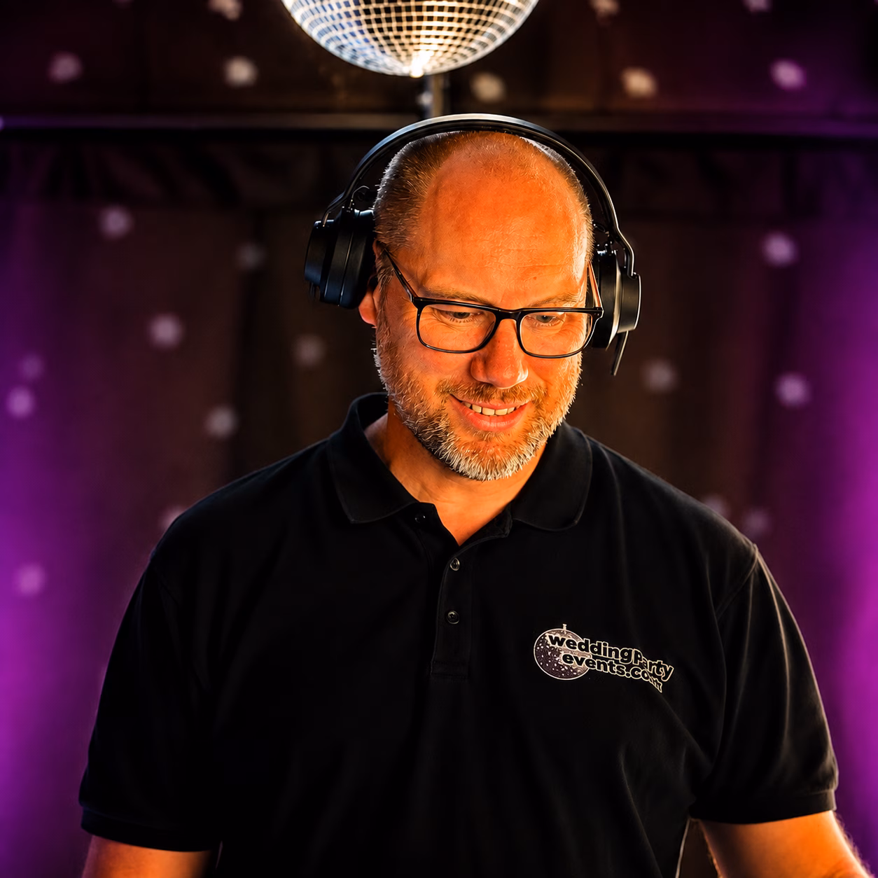 Wedding Party Events professional wedding DJ wearing headphones beneath a disco ball with purple uplighting.