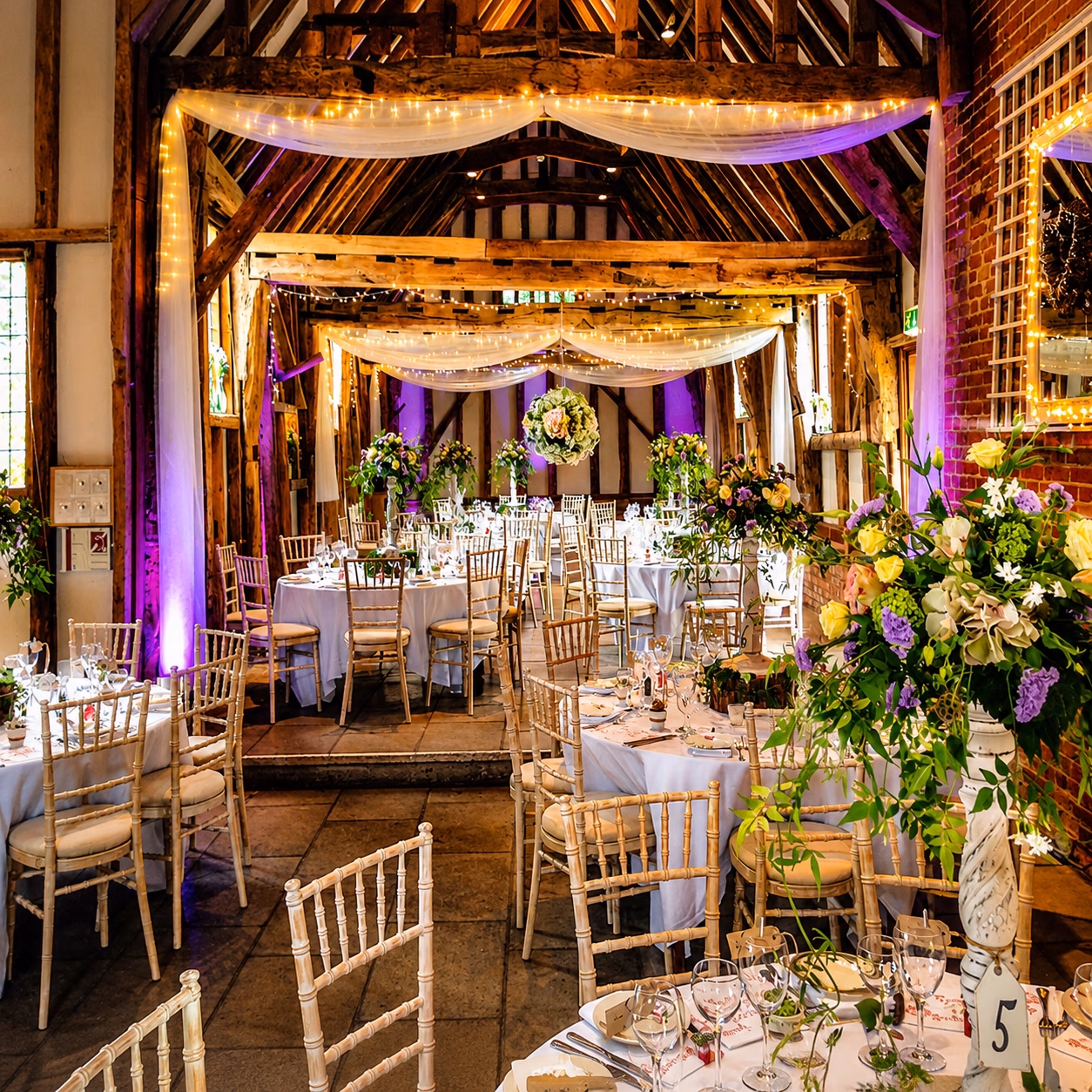 Elegant wedding reception setup at Haughley Park Barn in Suffolk, featuring rustic timber beams, floral centrepieces, fairy lights, purple uplighting and beautifully dressed wedding tables.